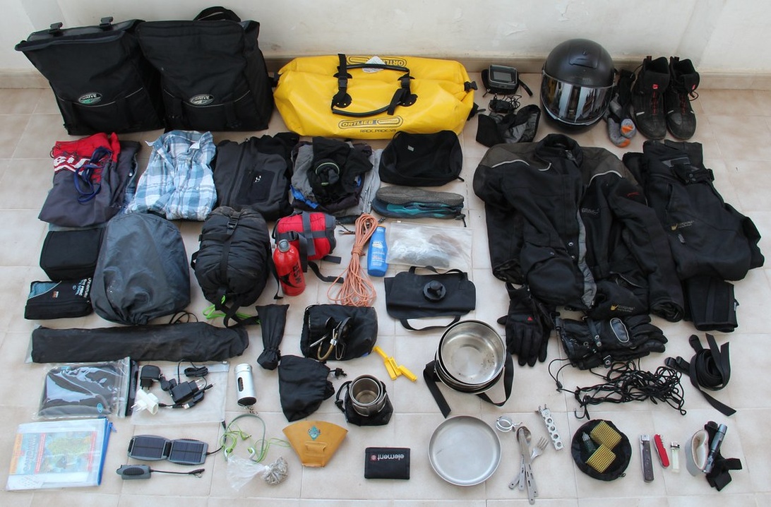 A flat lay of camping equipment including bags, clothing, electronic devices, and a helmet, illustrating the importance of organizing tech gear before a trip.